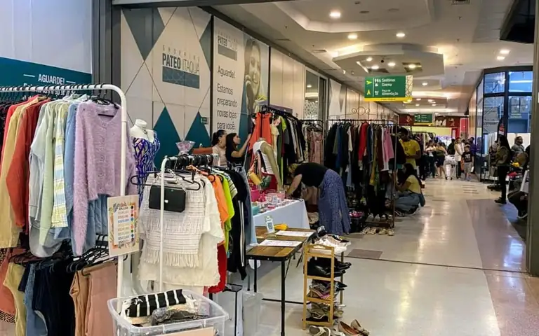 Corredor com expositores, roupas e visitantes em feira de brechó